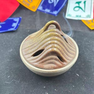 May include: A ceramic incense burner with a mountain-shaped lid, in brown and tan tones. The burner has a round base and several oval openings for smoke to escape. Wisps of smoke are visible, indicating use.