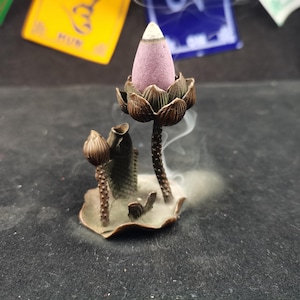 Puede incluir: Un quemador de incienso de color bronce en forma de flor de loto, con un cono de incienso púrpura que emite humo. El quemador tiene un diseño de pez y hoja. El fondo es oscuro.