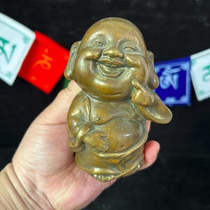 Könnte beinhalten: Eine bronzefarbene lachende Buddha-Figur. Die Statue hat einen großen Bauch, ein lächelndes Gesicht und hält den Finger hoch. Der Hintergrund umfasst farbenfrohe Gebetsfahnen.