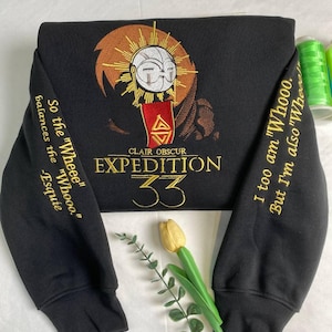 May include: Black sweatshirt with gold embroidery and a detailed graphic. The design includes the words "CLAIR OBSCUR EXPEDITION 33" and phrases on the sleeves. The graphic features a face with a halo and a red triangle.