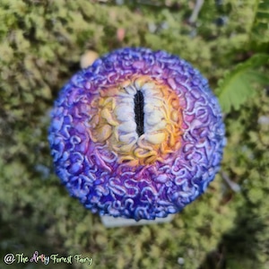 May include: A fantasy eye sculpture with a textured, multi-coloured design. The eye features shades of blue, purple, yellow, and orange, with a vertical black slit for the pupil. The sculpture is set against a mossy background.