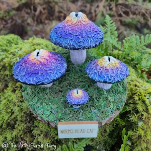 May include: A cluster of four Worms Head Cap mushrooms with textured, vibrant caps in purple, blue, and yellow hues. The mushrooms sit on a mossy base, with a wooden sign reading "WORMS HEAD CAP".