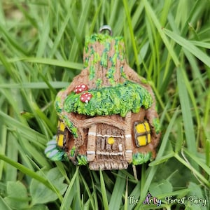 Pode incluir: Um pingente de casa de fadas em miniatura, feito com detalhes intrincados. A casa apresenta uma porta de madeira marrom, janelas iluminadas em amarelo e um telhado de musgo verde com cogumelos com manchas vermelhas. O texto "The Artsy Forest Fairy" é visível na parte inferior.