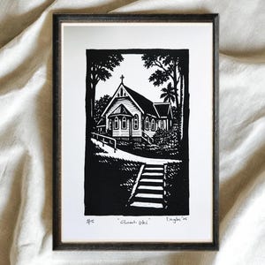 May include: Black and white framed print of a church, set in a wooded area. The artwork, titled "Church Uki," features a detailed linocut style with a cross atop the church. A set of stairs leads up to the building.