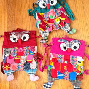 May include: Three colorful owl-shaped drawstring backpacks. The backpacks are made of patchwork fabric in red, pink, and teal. Each owl has large white eyes, a red beak, and fabric feet. The bags are on a wooden floor.