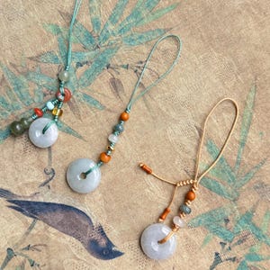 Puede incluir: Tres collares con colgantes de jade con piedras de jade circulares y cuentas de colores. Los colgantes están suspendidos de finos cordones. El fondo presenta una impresión de estilo vintage con bambú y un pájaro.