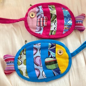 Puede incluir: Dos monederos hechos a mano con forma de pez. Uno es rosa con rayas verticales y un bot&oacute;n como ojo, el otro es azul con rayas verticales y un acento amarillo. Ambos tienen un lazo para llevar y una aleta.