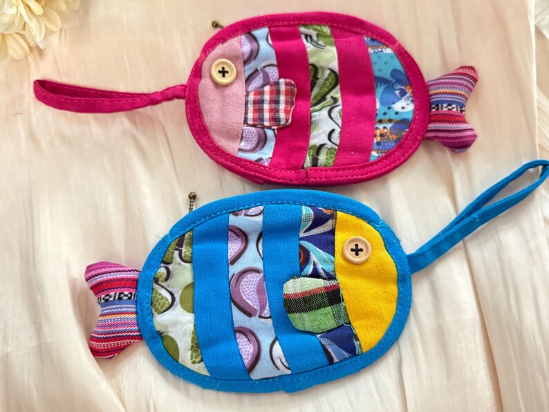 Puede incluir: Dos monederos hechos a mano con forma de pez. Uno es rosa con rayas verticales y un bot&oacute;n como ojo, el otro es azul con rayas verticales y un acento amarillo. Ambos tienen un lazo para llevar y una aleta.
