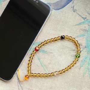 May include: A beaded phone charm with amber-colored beads and accents of red, green, and black beads. The charm is next to a black smartphone on a patterned surface.
