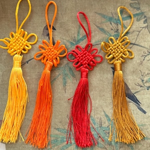May include: Four colorful Chinese knot tassels in shades of yellow, orange, and red. Each tassel features a decorative knot design at the top with a long, flowing fringe. The tassels are displayed against a patterned background.