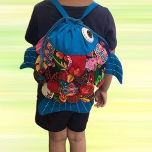 May include: A blue fish-shaped backpack with a drawstring closure. The backpack features colorful patchwork scales, blue fins, and a large eye. The straps are blue, and the bag is worn on the back.