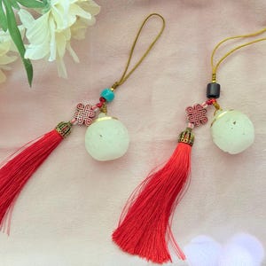 May include: Two decorative charms featuring long red tassels, gold-toned metal caps, and carved off-white spherical beads. Each charm includes a small red knotted accent, a gold-colored hanging loop, and a single bead in either turquoise or dark brown.