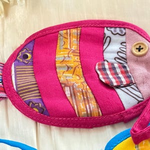 Puede incluir: Un art&iacute;culo de tela hecho a mano con forma de pez colorido. El pez es principalmente rosa intenso con secciones de tela estampada en morado, amarillo y blanco. Un bot&oacute;n de madera sirve como ojo y tiene un peque&ntilde;o lazo.