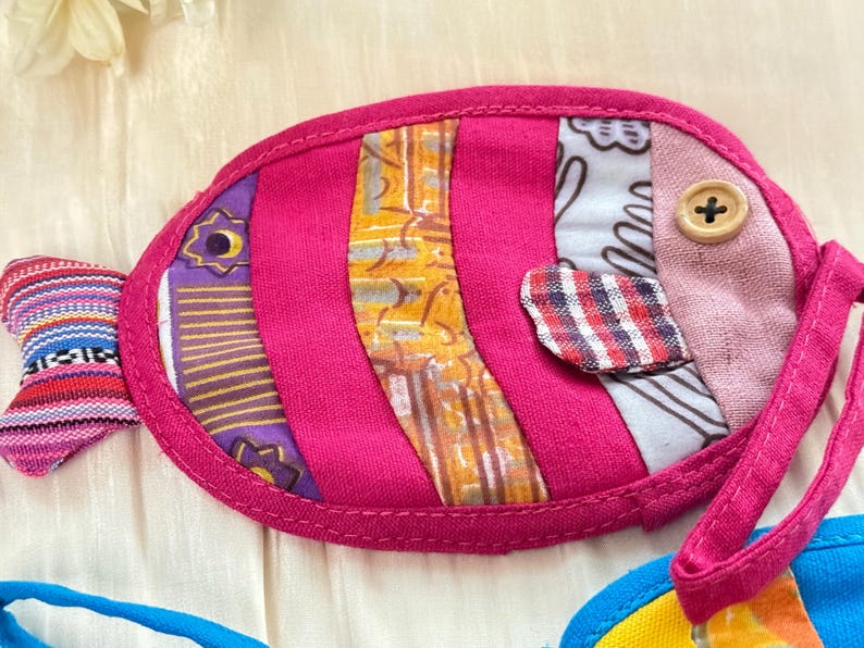 Puede incluir: Un art&iacute;culo de tela hecho a mano con forma de pez colorido. El pez es principalmente rosa intenso con secciones de tela estampada en morado, amarillo y blanco. Un bot&oacute;n de madera sirve como ojo y tiene un peque&ntilde;o lazo.