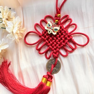 Puede incluir: Un colgante de nudo chino rojo con un dije decorativo de cabeza de león y una moneda de bronce. El colgante presenta un diseño tejido con una larga borla y detalles dorados. El fondo es una tela suave de color blanquecino.