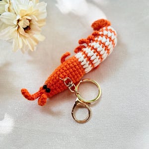 May include: A handmade orange and white crochet shrimp keychain with a gold-colored clasp and key ring. The shrimp has black eyes and small orange antennae. The keychain is a unique accessory.