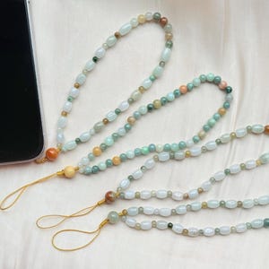May include: A close-up of three beaded phone charms. The charms feature a variety of pastel-colored beads, including white, green, and orange. Each charm has a gold-colored string and wooden bead detail.