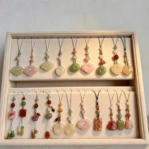 May include: A collection of decorative charms arranged in a beige display tray. Each charm features a cord with various stone beads, including jade, rose quartz, and carnelian, in shapes like circles, locks, and fruits, accented with small gold-toned metal beads.