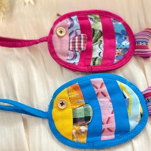 May include: Two handmade fish-shaped coin purses. One is pink with a button eye and striped accents, the other is blue with a yellow eye and patchwork details. Both have a loop handle and a striped tail.