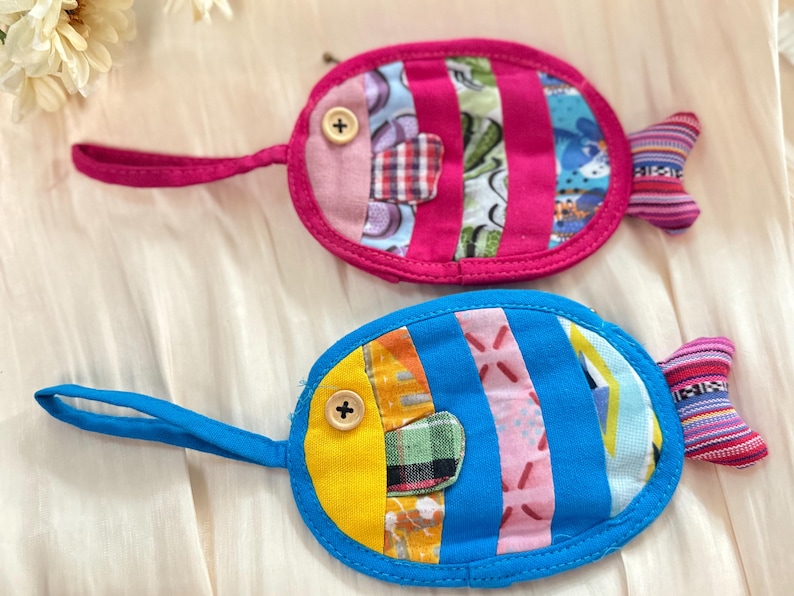 Puede incluir: Dos monederos hechos a mano con forma de pez. Uno es rosa con un ojo de bot&oacute;n y detalles a rayas, el otro es azul con un ojo amarillo y detalles de patchwork. Ambos tienen un asa de bucle y una cola a rayas.