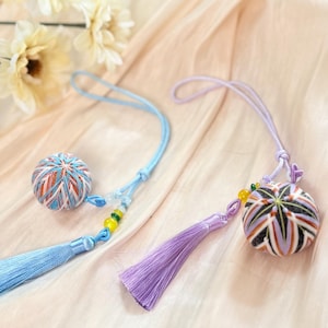 Könnte beinhalten: Zwei Temari-Bälle mit Quasten. Einer ist blau und orange, der andere lila und schwarz. Jeder hat eine passende Quaste und Kordel. Die Temari-Bälle liegen auf einem hellpfirsichfarbenen Stoff, mit einigen Blumen im Hintergrund.