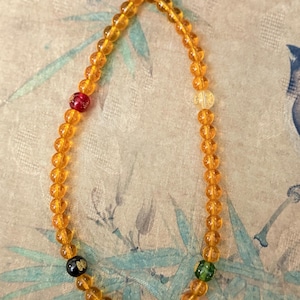 Może przedstawiać: Naszyjnik z koralików w kolorze bursztynu. Naszyjnik posiada akcentujące koraliki w kolorze czerwonym, żółtym, zielonym i czarnym. Jest nawleczony na żółty sznurek. Tło ma kwiatowy wzór.