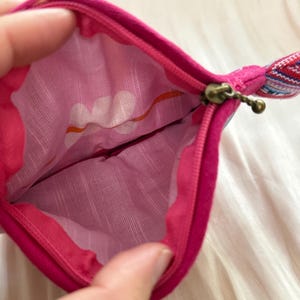 Puede incluir: Una bolsa con cremallera de color rosa intenso con un interior estampado. La tela interior presenta un dise&ntilde;o floral blanco con detalles en naranja. La cremallera es de color lat&oacute;n y la bolsa tiene un peque&ntilde;o lazo de tela con un estampado colorido.