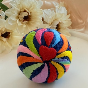 Puede incluir: Un cojín de alfileres redondo hecho a mano con una base azul marino, adornado con diseños bordados vibrantes. Los patrones coloridos incluyen un corazón rojo y arcos de colores del arcoíris. Un artículo decorativo y funcional para costureras.
