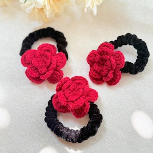 May include: Three black scrunchies with bright red, crocheted flower accents. The flowers are in full bloom, adding a pop of color against the dark bands. These hair accessories are perfect for adding a touch of handmade charm to any hairstyle.