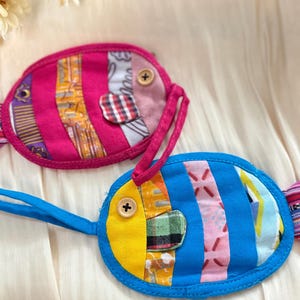 Puede incluir: Dos bolsas de tela hechas a mano con forma de pez. Una es rosa con un asa rosa, la otra es azul con un asa azul. Ambas tienen detalles de patchwork coloridos, ojos de bot&oacute;n de madera y aletas caudales estampadas. Estas bolsas probablemente se utilizan para objetos peque&ntilde;os.