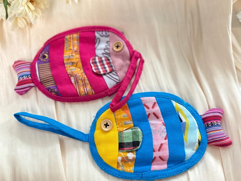 Puede incluir: Dos bolsas de tela hechas a mano con forma de pez. Una es rosa con un asa rosa, la otra es azul con un asa azul. Ambas tienen detalles de patchwork coloridos, ojos de bot&oacute;n de madera y aletas caudales estampadas. Estas bolsas probablemente se utilizan para objetos peque&ntilde;os.