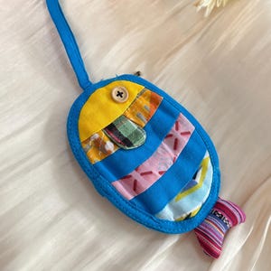 Puede incluir: Un llavero artesanal con forma de pez, con un lazo azul. El cuerpo es azul con parches de tela coloridos, una cabeza amarilla y una cola a rayas. Un bot&oacute;n de madera asegura la cabeza. El llavero mide aproximadamente 10 cm de largo.