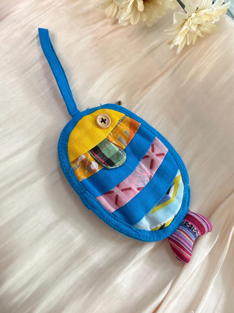 Puede incluir: Un llavero artesanal con forma de pez, con un lazo azul. El cuerpo es azul con parches de tela coloridos, una cabeza amarilla y una cola a rayas. Un bot&oacute;n de madera asegura la cabeza. El llavero mide aproximadamente 10 cm de largo.