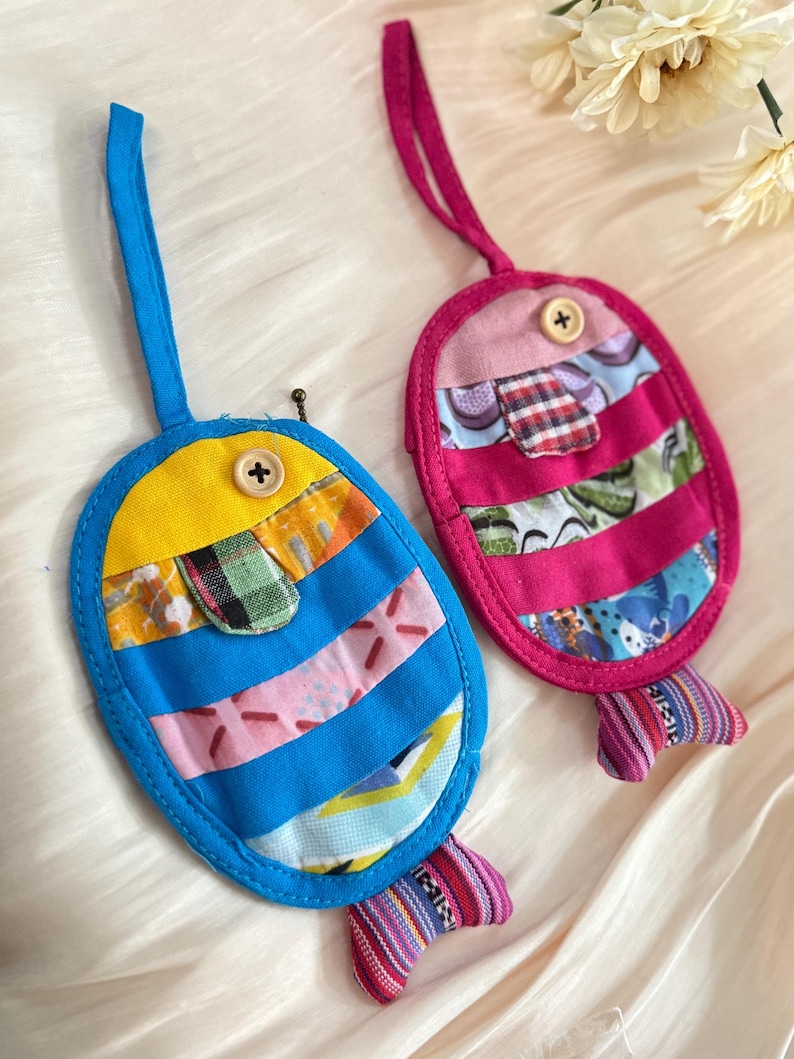 Puede incluir: Dos adornos artesanales de tela con forma de pez. Uno es azul y amarillo, el otro rosa. Ambos tienen cuerpos a rayas, detalles de botones y aletas caudales estampadas. Tienen un lazo para colgar.