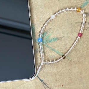 May include: A clear beaded phone charm with colorful accents, including blue, white, yellow, and red beads. The charm is attached to a black smartphone, resting on a patterned surface.
