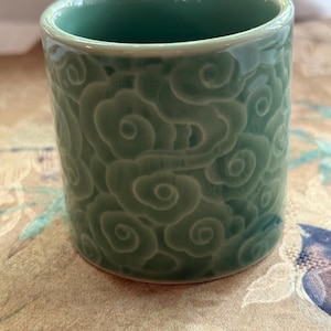 Puede incluir: Una taza de cerámica verde celadón con forma cilíndrica. El exterior de la taza presenta un patrón de nubes repetido. La taza está sobre una tela estampada.