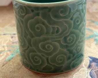 May include: A celadon green ceramic cup with a cylindrical shape. The cup's exterior features a repeating cloud pattern. The cup is sitting on a patterned fabric.
