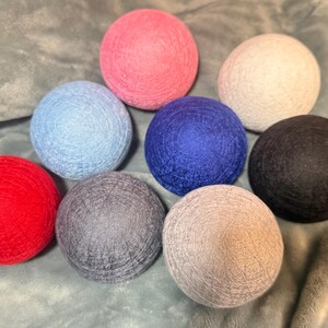 May include: A collection of eight spherical decorative balls wrapped in textured thread, featuring a vibrant color palette of red, light blue, pink, dark blue, white, black, grey, and beige, arranged in a cluster on a soft fabric surface.