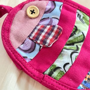 Puede incluir: Una m&aacute;scara para los ojos hecha a mano con forma de pez. Tiene un borde rosa brillante y est&aacute; hecha con retales de tela en tonos rosa, azul y verde. Un peque&ntilde;o bot&oacute;n beige est&aacute; cosido como elemento decorativo.