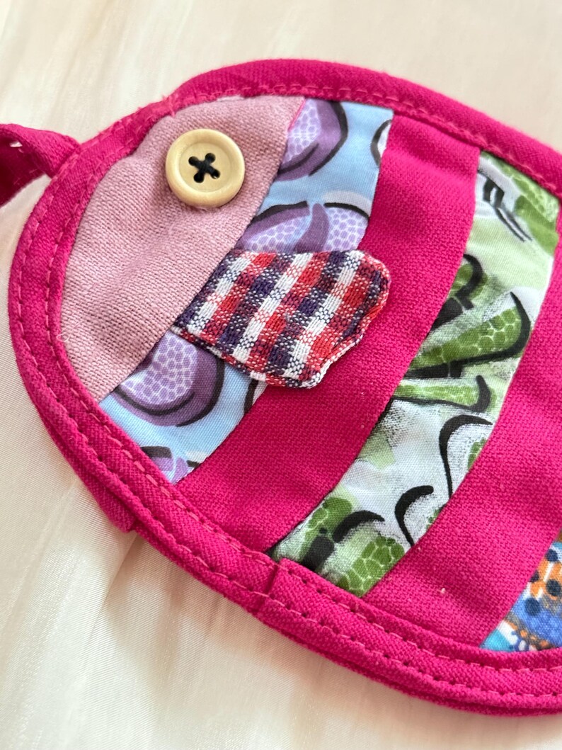 Puede incluir: Una m&aacute;scara para los ojos hecha a mano con forma de pez. Tiene un borde rosa brillante y est&aacute; hecha con retales de tela en tonos rosa, azul y verde. Un peque&ntilde;o bot&oacute;n beige est&aacute; cosido como elemento decorativo.