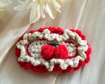 Puede incluir: Pinza para el pelo hecha a mano con ganchillo, con un diseño rojo y blanco y un motivo de cereza. La pinza tiene un borde festoneado y un detalle de tallo verde. El fondo es de tela suave y neutra.