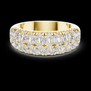 May include: A wide, gold-colored ring with three rows of sparkling, clear gemstones. The ring has a smooth, polished band and is set against a black background. The gemstones are round-cut and closely set together, creating a continuous sparkle.