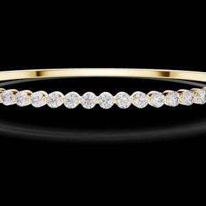 May include: A gold bangle bracelet with a row of round, clear gemstones. The bracelet is set against a black background, highlighting the sparkle of the gemstones and the shine of the gold.