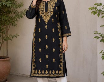 May include: Black tunic with gold embroidery. The long-sleeved tunic features intricate gold patterns around the neckline, down the front, and on the sleeves. The tunic is paired with white trousers and brown flats. The outfit is suitable for casual or semi-formal occasions.