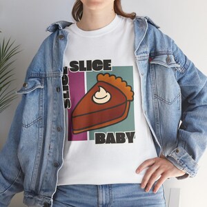 Könnte beinhalten: Weißes T-Shirt mit einem Tortenstück-Motiv und Sahne, sowie dem schwarzen Schriftzug "SLICE BABY". Die Person trägt eine blaue Jeansjacke.