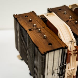 May include: Close-up of a computer CPU cooler with a wooden top and metal fins. The cooler has a brown wooden frame with the brand name "Noctua" engraved on it. The fins are silver and tightly packed.