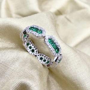 Puede incluir: Un anillo de tono plateado que presenta una serie de eslabones rectangulares, cada uno engastado con una fila central de pequeñas piedras preciosas de color verde vibrante rodeadas por un halo de diminutas piedras blancas brillantes.