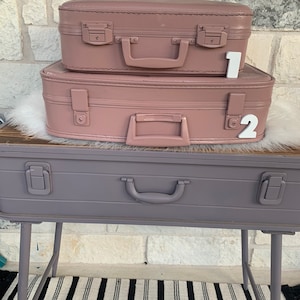 May include: Two vintage suitcases stacked on top of each other. The suitcases are painted a dusty rose color and have metal clasps and handles. The suitcases are sitting on a purple suitcase table with metal legs. The table is sitting on a black and white striped rug.