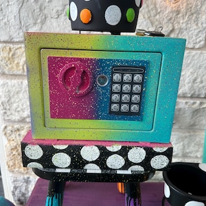 May include: A colorful, hand-painted safe with a combination lock, set atop a stool. The safe is painted with a rainbow gradient and a black and white polka dot pattern. The stool legs are painted in various colors with paint drips.