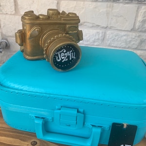 May include: A gold-colored camera figurine sits on top of a teal blue suitcase with a black tag labeled "17". The camera lens has a black circle with white lettering that says "Jez4u".